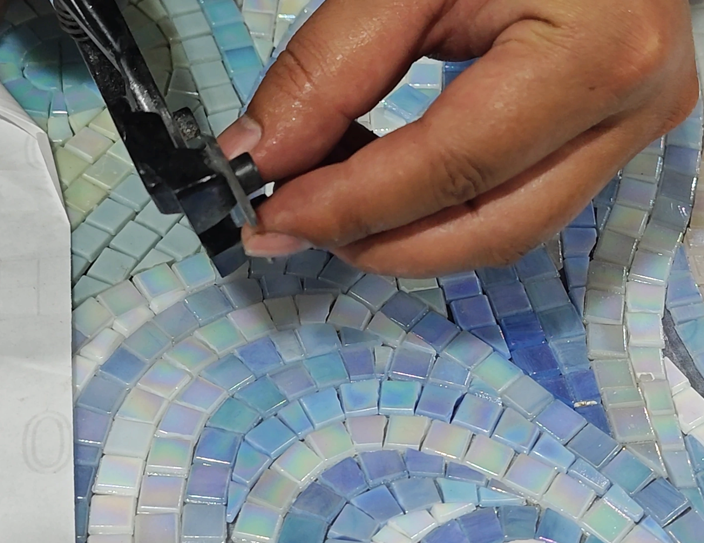 Artisan using mosaic nippers to shape a small tile over a curved mosaic pattern